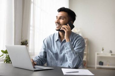Merkez büroda dizüstü bilgisayarla çalışan pozitif Hintli bir adam, müşteriye profesyonel danışmanlık sağlıyor cep telefonu kullanıyor, resmi konuşmaları yönetiyor, işi telefonla çözüyor, iletişimden keyif alıyor.