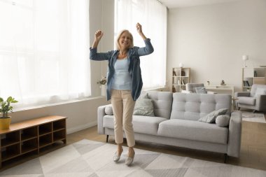 Neşeli, yaşlı ve emekli bir kadın, oturma odasında dans ediyor, zıplıyor, galibiyeti kutluyor, başarıyı kutluyor, enerjik hareketten, müzikten, modern apartmanın içinde boş vakitlerden keyif alıyor.