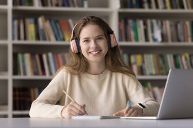 Güzel 16 'lı yaşlardaki kız üniversite sınavlarına hazırlanıyor ya da üniversiteye kabul ediliyor modern teknolojiyi kullanıyor, kütüphanede masa başında oturuyor, kulaklık takıyor kameraya bakıyor. Mükemmel bir lise öğrencisinin portresi, kendini yetiştirme.
