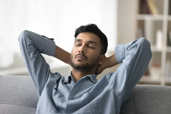 stock image Indian man take break leaned on soft couch in living room, relaxing, settled down on couch with hands behind head, felt asleep, having healthy daytime nap alone at home. Stress-free, reposing, pause