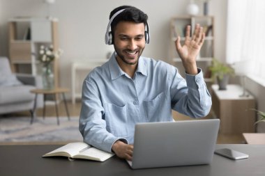 Masada oturan genç Hintli adam kablosuz kulaklık takıyor öğretmenini selamlıyor yeni bilgi edinmek, dil öğrenmek, çevrimiçi çalışmak, dizüstü bilgisayar ve modern video konferans uygulaması kullanarak video dersine başlıyor.