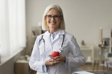 Göğsünde kırmızı kalp şeklinde bir nesne tutan mutlu kıdemli doktor kadın dişlek gülüşüyle kameraya bakıyor. Olgun bir pratisyen, beyaz üniformalı ve gözlüklü bir kardiyolog.