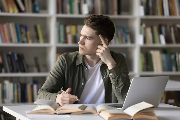 stock image Pensive student guy stuck on difficult task for hours without progress, staring aside feels annoyed looks confused sit at desk with textbooks and laptop. Hard exercise, lack of skills or understanding