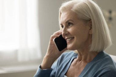 Evde cep telefonuyla konuşan, gülümseyen, gülen, sohbetten zevk alan, modern telsiz iletişimi olan yaşlı, pozitif sarışın bir kadın. Yan görünüş, sıradan bir portre.
