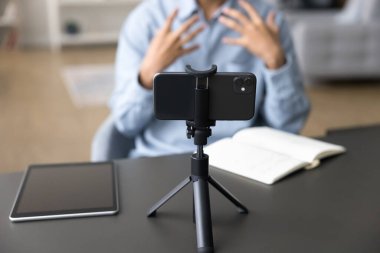 Cep telefonunun tripoda durmasına odaklanın, arka plandaki erkek vlogger konuşmasına, yeni video kaydına veya akıllı telefon kamerasına eğitici webinar kaydına. Çevrimiçi yayın, sosyal medya simgesi, etkileme, blog yazma