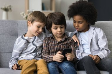 Üç çocuk birlikte akıllı telefon kullanıyor, kanepede oturuyor, evde boş zaman geçiriyor, cihazla eğleniyor, çevrimiçi yeni oyunlar oynuyor, arkadaş canlısı kardeşler sosyal ağda komik videolar izliyor, internette geziniyor.