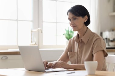 Evde çalışan ciddi orta yaşlı bir kadın, dizüstü bilgisayar kullanıyor, rapor hazırlıyor, e-banka başvurusuyla online ödeme yapıyor, rahat yerli mutfağındaki masada oturuyor. Telework, modern teknoloji kullanımı