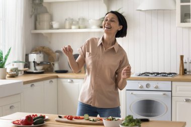 Neşeli orta yaşlı kadın modern mutfakta dans ediyor, yemek yapıyor, sağlıklı yemek hazırlıyor, sebze salatası kesiyor, eğleniyor, yemek pişiriyor ve evde vakit geçiriyor. Mutfak, hobi, yaşam tarzı
