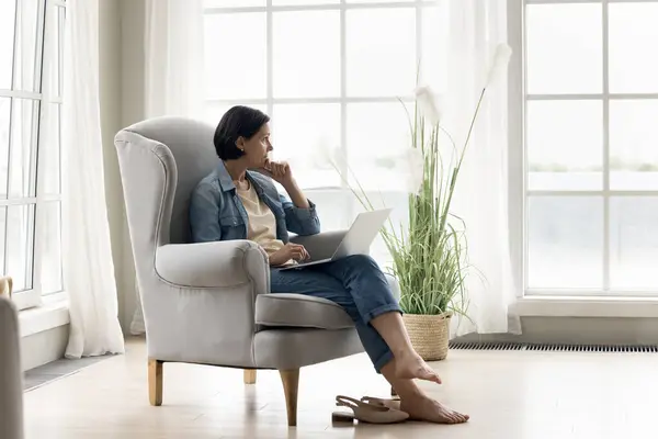 stock image Pensive mature woman working on laptop at home, sitting on armchair with computer on laps, staring into distance, thinking, search problem solution, ponder bad on-line news feels concerned or fatigued