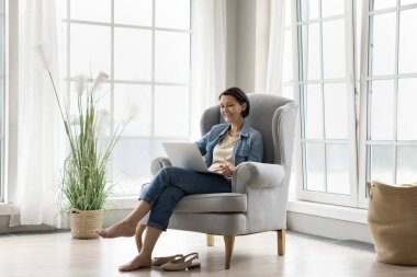 Neşeli orta yaşlı kadın rahat koltukta rahatlıyor dizüstü bilgisayara bakıyor komedi filmi izlerken gülüyor, yetişkin çocuklarla uzaktan iletişim kuruyor video çağrısı uygulaması kullanıyor.