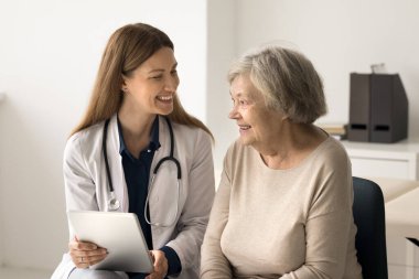 Üniformalı gülümseyen genç bayan doktor dijital tablet taşıyor. Tıbbi muayene sonucu, muayene ve elektronik reçeteyi açıklıyor. Yaşlı hasta profesyonel yardım alıyor