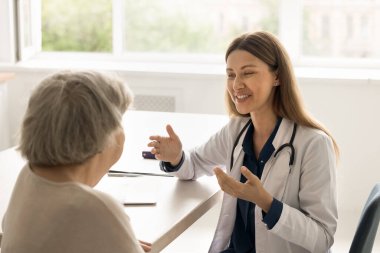 Üniformalı kadın doktor, masa başında oturmuş yaşlı kadın hastayla iletişim kuruyor, tedaviyi açıklıyor, tavsiye veriyor. Klinik ziyareti sırasında yaşlı bir kadın konsültasyon alıyor. Geriatri, sağlık hizmetleri