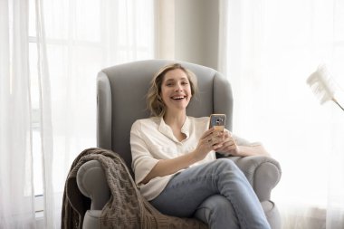 Gülümseyen genç bir kadın cep telefonu tutuyor, gülümsüyor, kameraya bakıyor, rahat koltukta rahatlıyor, modern teknolojiyi kullanıyor, internette vakit geçiriyor, telefonla konuşuyor, çevrimiçi arkadaşlarla iletişim kuruyor.