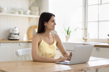 Gülümseyen kadın mutfakta dizüstü bilgisayarla masada oturuyor, sipariş veriyor, online süpermarketten ürünler alıyor, modern bilgisayar kullanarak çalışıyor ya da çalışıyor, evde internetin keyfini çıkarıyor.