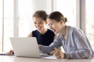 İki genç, mutlu ofis çalışanı, kadın meslektaşlar, takım arkadaşları dizüstü bilgisayarla masaya oturur, konuşurlar, işbirliği projesinde birlikte çalışırlar, fikirleri paylaşırlar, başarılı bir takım çalışması yaparlar, iş yerinde eğlenirler.