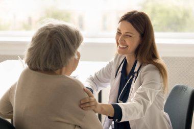 Üniformalı genç bayan pratisyen doktor omuzlarına dokundu, destek verdi ve profesyonel tıbbi bakım yaptı. Yaşlı kadın klinikteki randevuda doktorla konuşuyor. Geriatri, kontrol.