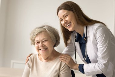 Gülümseyen genç bayan doktor klinikteki mutlu yaşlı kadının omuzlarına sarılıyor, konsültasyon sırasında destek oluyor, psikolojik yardım sağlıyor. Kıdemli hasta iyi tıbbi hizmetlerden hoşlanır. İlaç, yaşlı bakımı.
