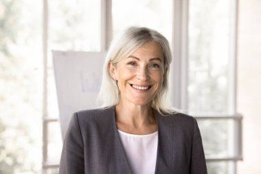 Resmi kıyafetli güzel orta yaşlı iş kadını gülümseyip modern ofiste yalnız poz veren kameraya bakıyor. Profesyonel işçi, satıcı, İnsan Kaynakları müdürü ya da işletme eğitmeni, kapalı alan portresi.