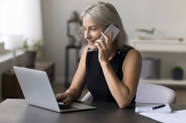 Çekici gülümseyen iş kadını modern ofiste masa başında oturur, akıllı telefon tutar, iş görüşmesi yapar, hoş sohbetin tadını çıkarır, müşteriye telefonla danışır, ortaklarla proje hakkında konuşur, dizüstü bilgisayar kullanır.