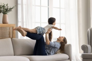 Sevgi dolu anne, tatlı kızıyla kanepede uzanıyor, onu kollarını açıp kaldırıyor, evde birlikte oyun oynuyor, hafta sonları rahat bir oturma odasında eğleniyor.