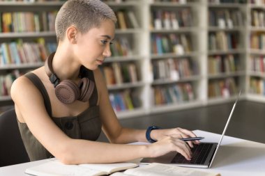 Üniversite kütüphanesinde okuyan kısa saçlı bir kız öğrenci dizüstü bilgisayarda klavye kullanıyor, kompozisyon hazırlıyor, özet yapıyor, modern teknoloji kullanarak tez üzerinde çalışıyor, okul ödevleri için internette arama yapıyor.