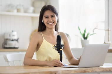 Genç bayan blogcu kablosuz kulaklık takıyor mutfaktaki masada dizüstü bilgisayar ve mikrofon tebessümüyle kameraya bakıyor. Sosyal medya simgesi, etkileyici portre, blog, çevrimiçi öğretim, e-koçluk