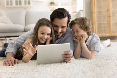 Gülümseyen baba evde küçük çocuklarla vakit geçiriyor, arkadaş canlısı aile oturma odasında uzanıyor dijital tablet kullanıyor, komik videolar seçiyor, akrabalarla uzaktan sohbet ediyor, internetten alışveriş yapıyor.