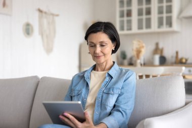 Gülümseyen orta yaşlı bir kadın dijital tabletle kanepeye oturur, kablosuz interneti tarar, evdeki koltukta rahatlar, ped cihazı kullanır, serbest çalışır, uzaktan sohbet eder, e-banka uygulaması üzerinden dengeyi kontrol eder.