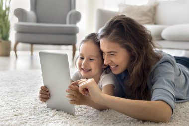 Mutlu genç anne evde küçük kızıyla vakit geçiriyor, yerde uzanıyor birlikte dijital tablet kullanıyor, çizgi film izliyor, sosyal ağlarda komik videolar izliyor, modern uygulamaların tadını çıkarıyor.
