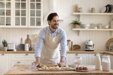 Yakışıklı gülümseyen adam bisküviler için önlük takar ya da rulo raptiyeli ev yapımı hamur tatlısı, yemek masasında durur, mutfakta tek başına vakit geçirir, yemek pişirmekten zevk alır. Hobi, ev işleri, yemek tarifi.