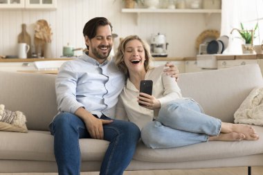 Gülümseyen çift oturma odasında kanepede oturur, cep telefonuyla vakit geçirir, ekran cihazına bakar, arkadaşlarıyla görüntülü sohbet eder, çevrimiçi komik video içeriği izler, modern teknolojiyi kullanarak eğlenir.