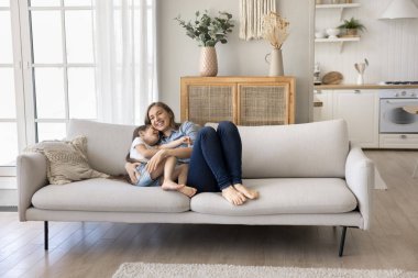 Sevgi dolu anne ve küçük kızın tatlı anları. Sevimli anaokulu çocuğu kanepede dinleniyor sevgili annesine sarılıyor, gülüyor, oynuyor, kanepede oturuyor evdeki rahat oturma odasında kaygısızca dinleniyor.