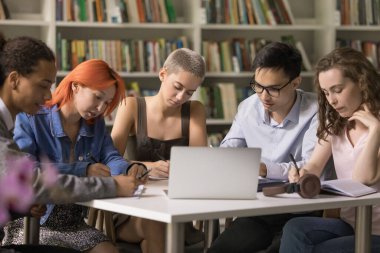 Çok ırklı bir grup öğrenci kütüphanede masalarında oturuyor, not yazıyor, defterde yazıyor, ödev yapıyor veya makale yazıyor, ders veya eğitim semineri sırasında öğrenim ve kişisel gelişim ile uğraşıyor.