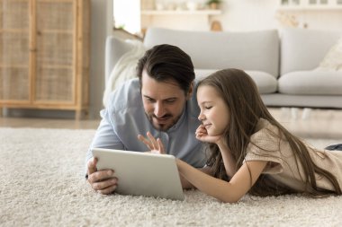 Önemseyen genç baba, sevimli küçük anaokulu kızıyla birlikte yerde yatıyor, dijital tablet ekranlara bakıyor, internetten alışveriş yapıyor, çizgi film izliyor, internet içeriğinin keyfini çıkarıyor.