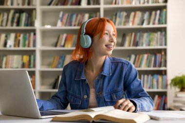 Kızıl kafalı kız öğrenci ses derslerini çevrimiçi okuyarak lise kütüphanesinde dizüstü bilgisayar ve ders kitaplarıyla oturuyor, üniversiteye giriş için hazırlanıyor, öğrenme sürecinin keyfini çıkarıyor, internet kaynakları kullanıyor.