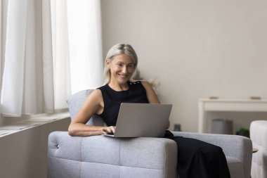 Gülümseyen yetişkin bir kadın oturma odasında dizüstü bilgisayarla oturur, online servis kullanır, internetten alışveriş yapar, serbest çalışan bir kadın uzaktan evde çalışır, e-postaları kontrol eder. Modern teknoloji