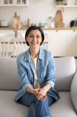 Attractive middle-aged woman in casual clothes smile look at camera relaxing sit on cozy sofa in studio apartment feels happy, enjoy pleasant weekend at own or rented flat. carefree retiree portrait clipart