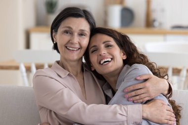 Neşeli, olgun 50 'li yaşlarda bir anne, yetişkin bir kıza kafa teması ile sarılıyor, koltukta dinleniyor, kameraya bakıyor, gülümsüyor, gülüyor, yakınlık kuruyor, aile bağı kuruyor, annelik yapıyor.