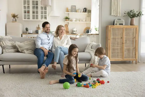 stock image Smiling carefree parents spend weekend time with cute little 6s children at home, look how siblings play colorful constructor game seated on warm floor. Pastime of friendly young family with two kids