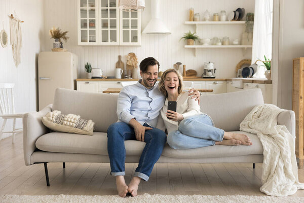 Happy young couple spend weekend in living room, hold smartphone sitting on couch, look at screen make selfie on mobile phone, smile, hug, watch funny internet content, enjoy remotely communication