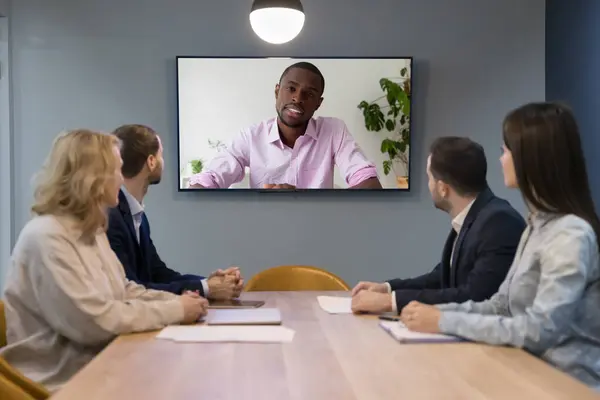 stock image Group of businesspeople take part in videoconference sit at desk look at tv monitor, African businessman, team leader or boss lead virtual meeting with staff or clients, make speech, share information