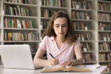 Güzel 18 'lik öğrenci kız yeni bilgiler ediniyor, üniversite kütüphanesinde eğitim görüyor, dizüstü bilgisayarla masaya oturuyor, ders kitaplarından başlıklar okuyor, notlar alıyor, özet yazıyor. Yüksek öğrenim, kalkınma