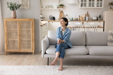 Modern oturma odasındaki kanepede dinlenen mutlu, güzel bir kadın. Uzağa bakıp gülüyor, kendi modern dairende kaygısız rahatlığın tadını çıkarıyor. Sorunsuz yetişkin dişiler evde yalnız vakit geçirir.