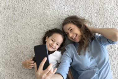 Mutlu anne ve kız yerde uzanıp cep telefonu ekranına bakıyorlar, selfie çekiyorlar, cep telefonu uygulamasını kullanarak eğleniyorlar, internette boş zaman geçiriyorlar, video konferansı yapıyorlar, üstten bakıyorlar.