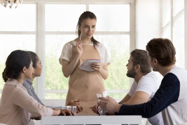 Önlük üniformalı olumlu garson not defteri tutuyor, müşterilere servis yapıyor, müşterilere menüyü tavsiye ediyor, notlar yazıyor. Bir grup genç insan, neşeli çokuluslu kızlar ve erkekler modern bir yerde yemek yemeye geldiler.