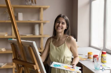 Çekici ressam elinde paletle poz veriyor ve resim fırçası sehpası üzerinde tuvalin yanında duruyor gülümsüyor kameraya bak fotoğraf çekimlerinin keyfini çıkar, sanat sınıfı atölyesinde yaratıcı bir hobi. Kadın ressamın portresi