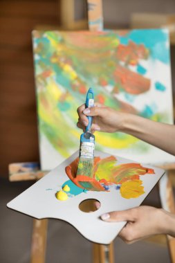 Dikey yakın görünüm bilinmeyen kadın ellerinde palet ve boya fırçası, akrilik boya renkleri karıştırma resim çizme, profesyonel sanat atölyesinde tek başına hobi veya profesyonel mesleğin keyfini çıkarma