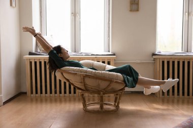 Modern kase şeklindeki sandalyede dinlenen mutlu 25 'li yaşlarındaki kadın kollarını kaldırıyor tatmin oluyor, hafta sonu boş zamanlarının keyfini çıkarıyor sıcak oturma odasında, yan bakışta. Modern mobilya, kiracılık, dinlenme