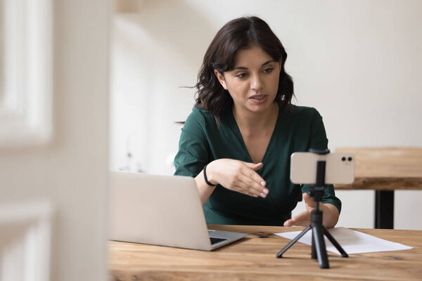 Деловая женщина тысячелетия, влиятельная запись нового видео-блога на камеру смартфона, сидит за столом домашнего офиса. Женщина-блоггер снимает видеоблог, ведет прямую трансляцию с последователями, общается он-лайн