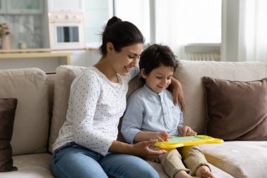 Gülümseyen Hintli anne ve oğlu renkli mıknatıs çizim tahtasıyla oynuyorlar, küçük anne ve 5 'li yaşlardaki çocuklara sarılıyorlar, kanepede oturuyorlar, hafta sonlarını evde eğitim oyuncaklarıyla geçiriyorlar.